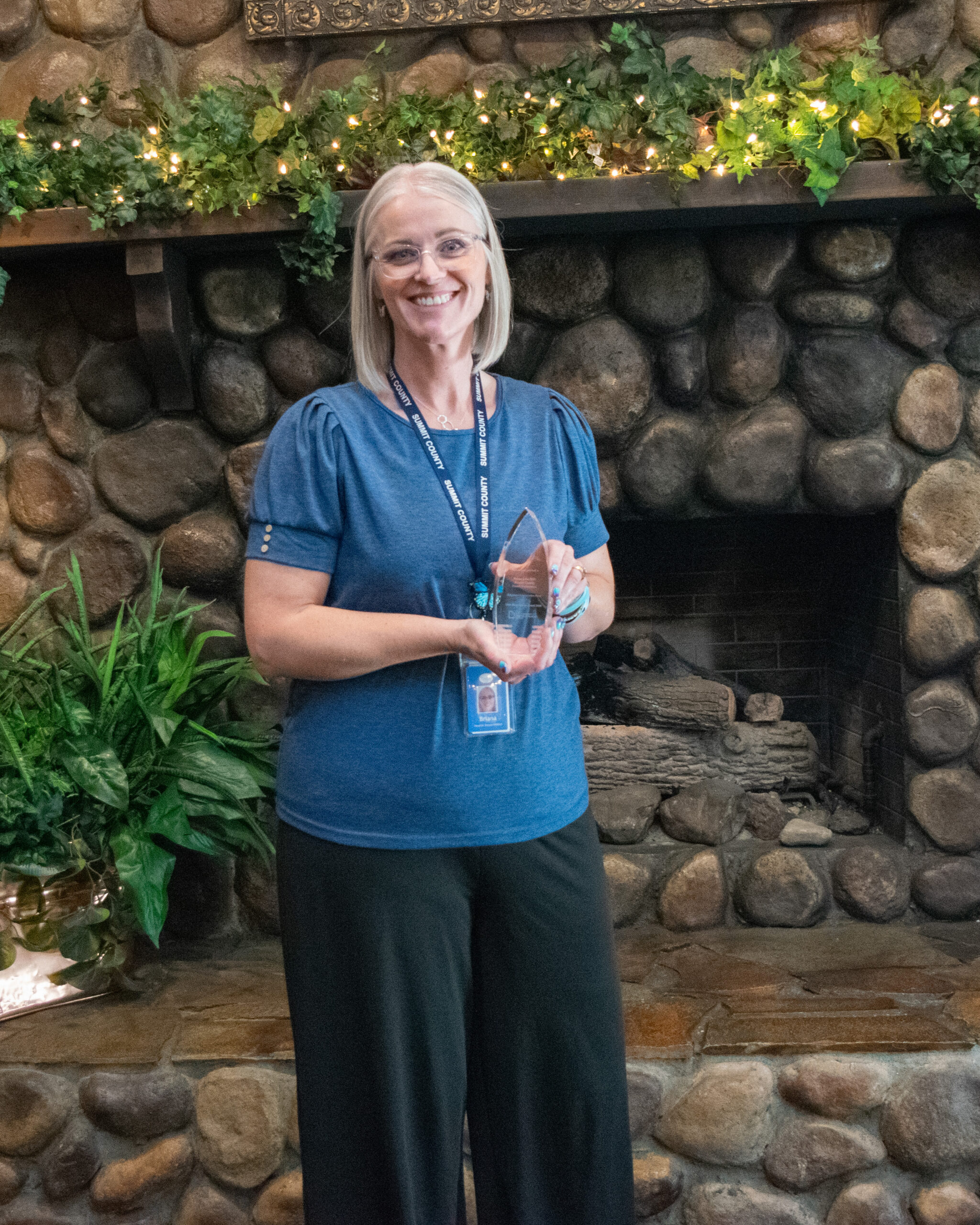Briana Jones in a blue shirt holding a Silver syringe award, standing in front of a stone fireplace.