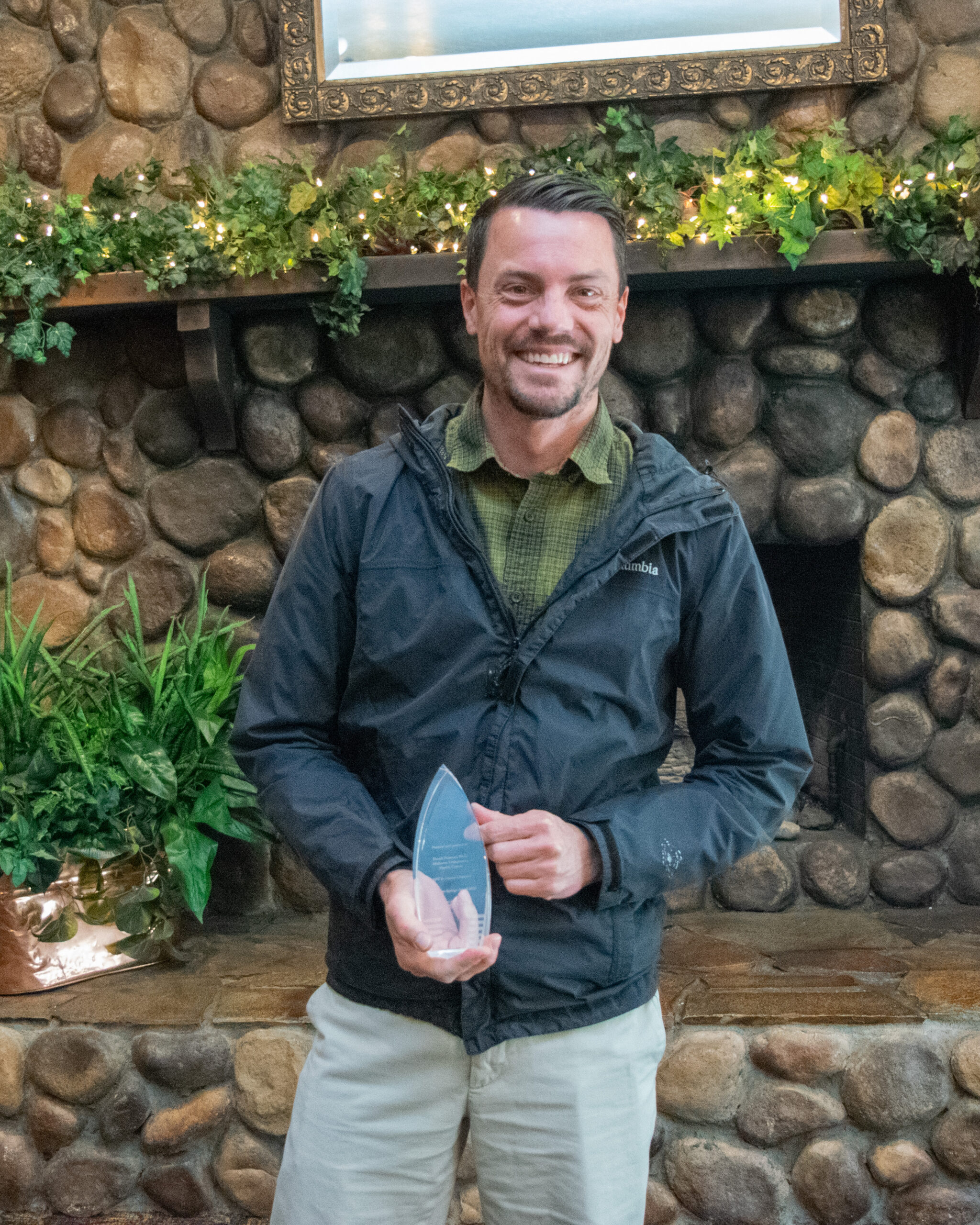 Derek Peterson, PA-C holding a Silver syringe award stands in front of a stone fireplace with plants and lights.