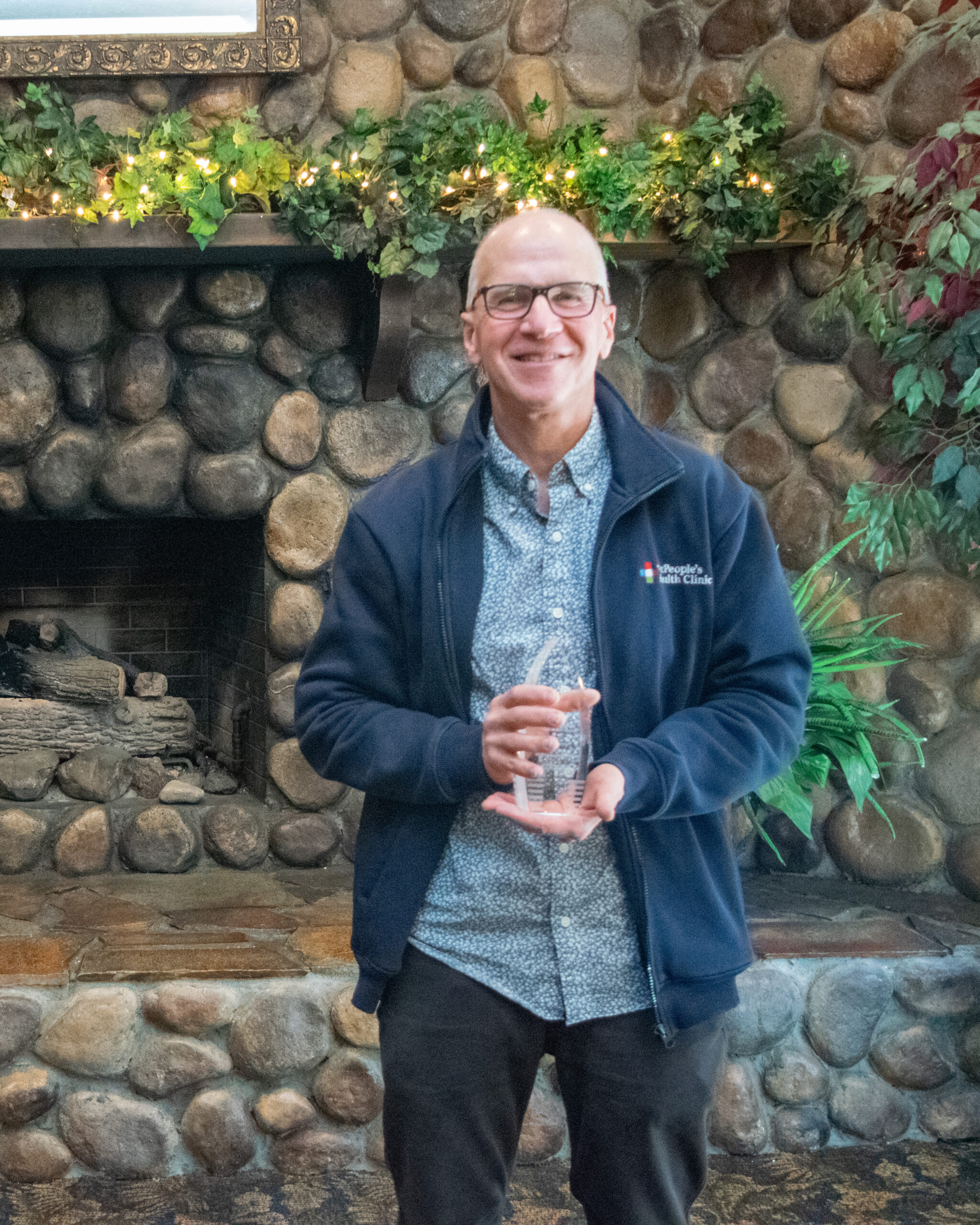 Scott Shulman holding a Silver syringe award in front of a stone fireplace with greenery overhead.