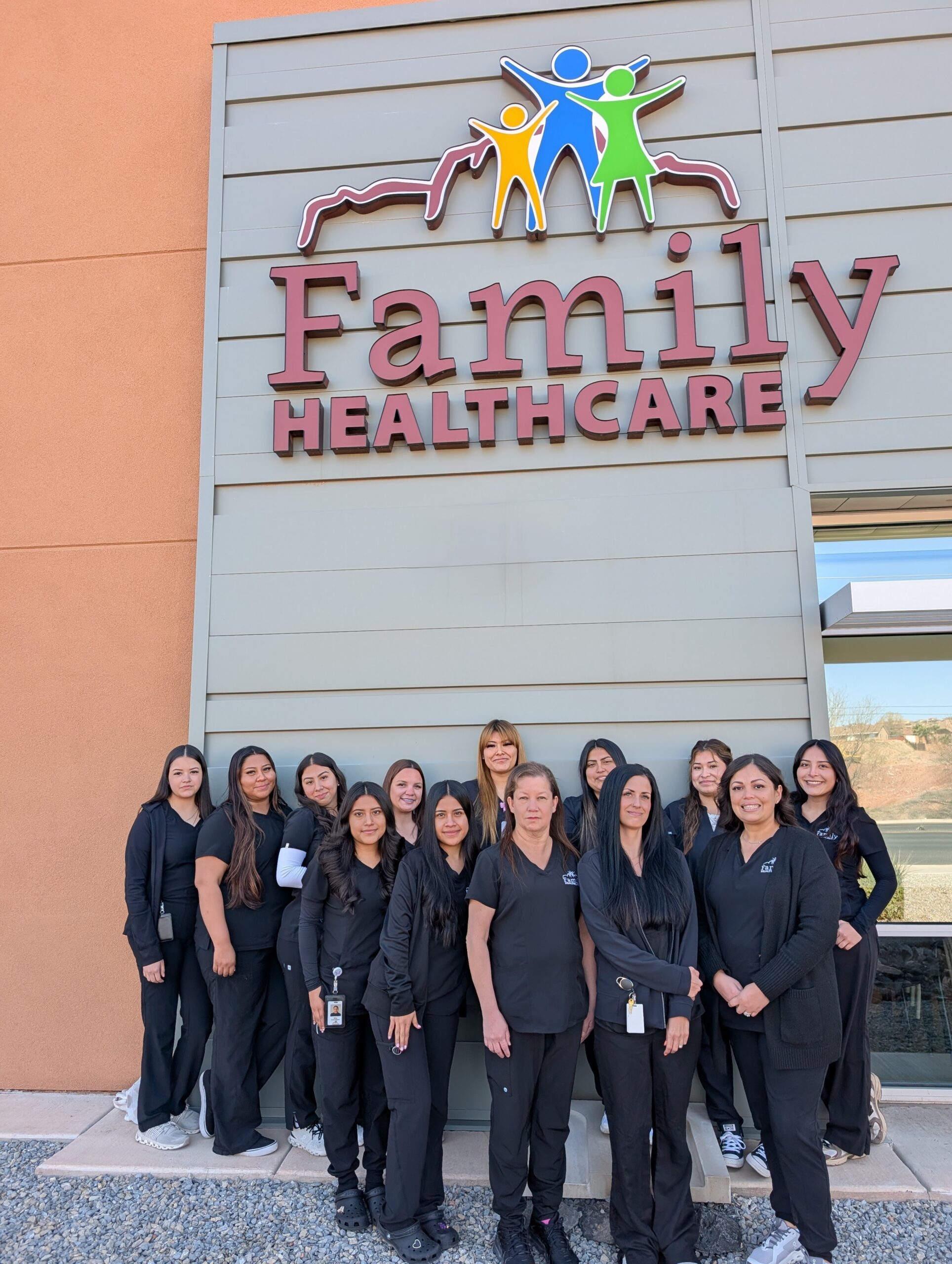 Twelve women in black uniforms stand smiling by a Family Healthcare sign on a building.