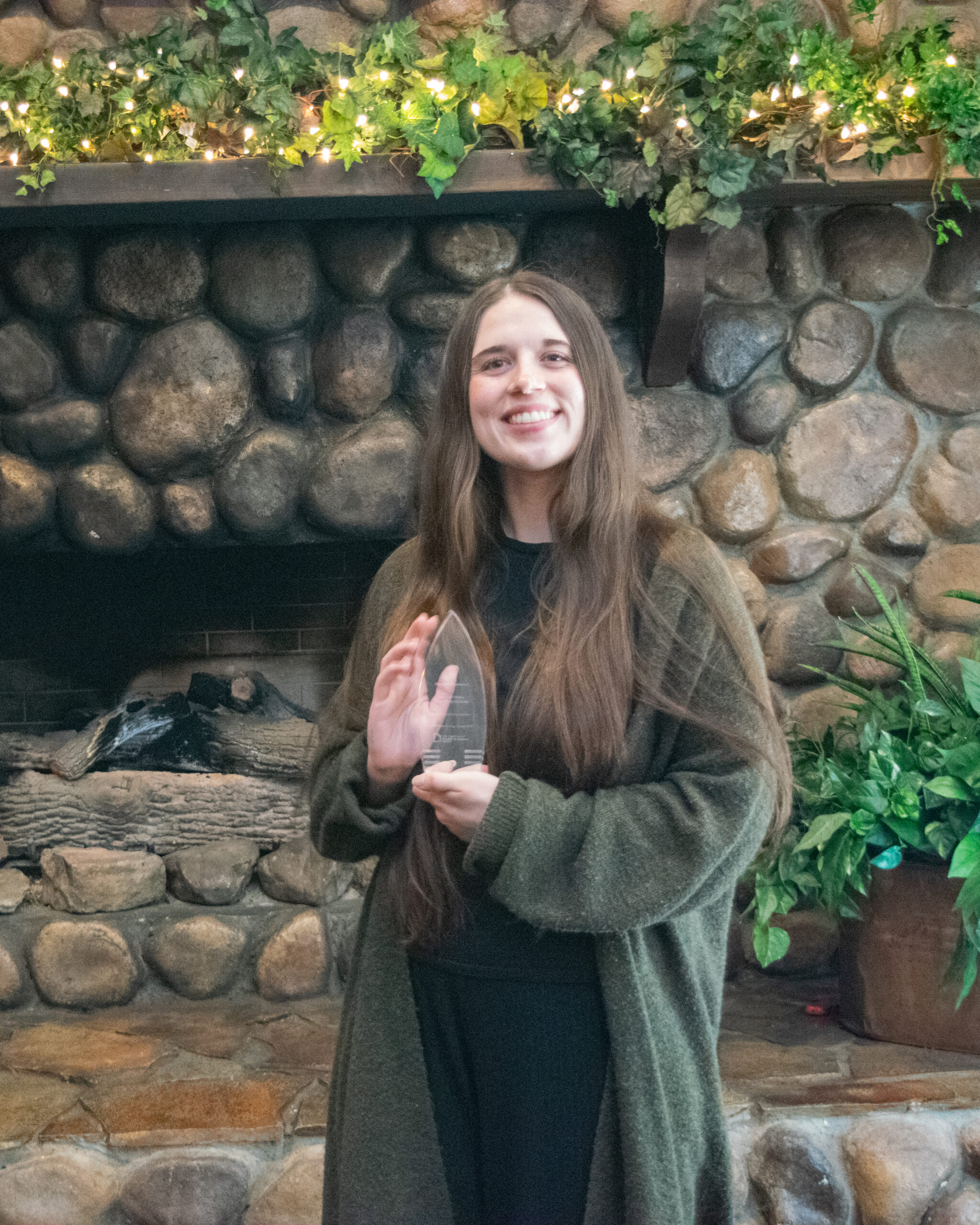 Gabrielle Mojica smiling and holding a Silver syringe award in front of a stone fireplace with plants and lights.