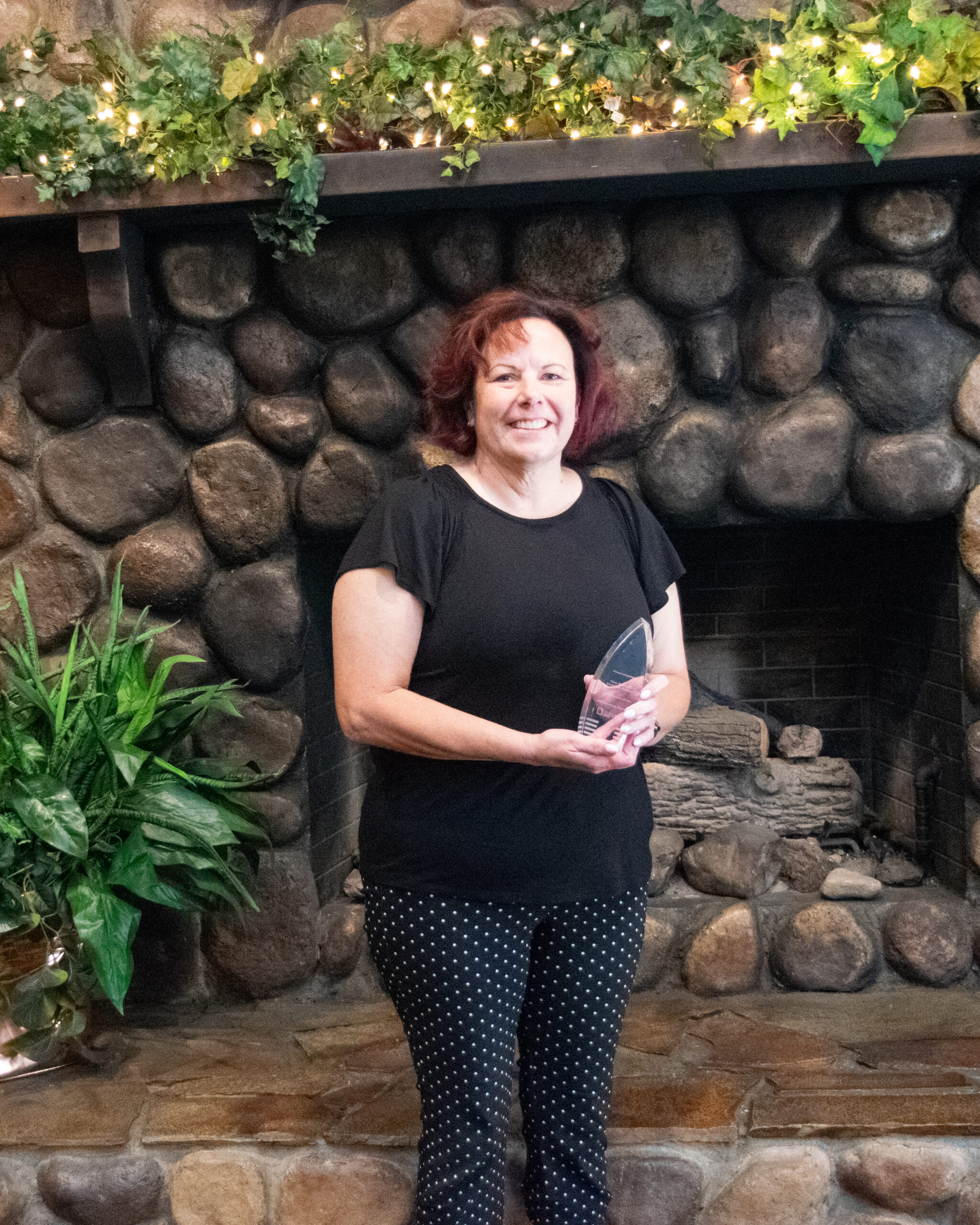 Ginny Murphy holding a Silver syringe award stands in front of a stone fireplace with greenery and lights above.