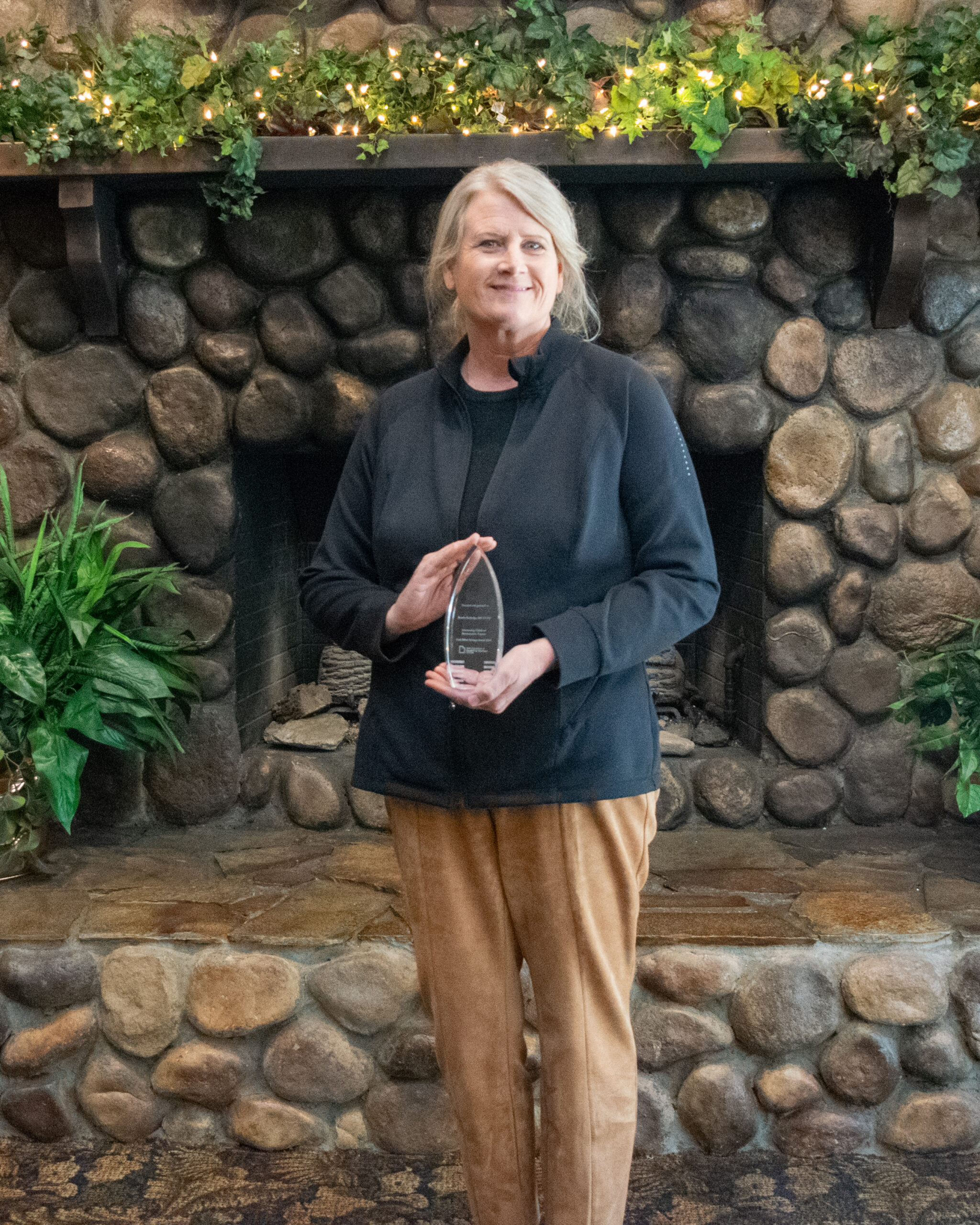 Sandy Rutledge holding the Silver Syringe award stands in front of a stone fireplace decorated with plants and lights.