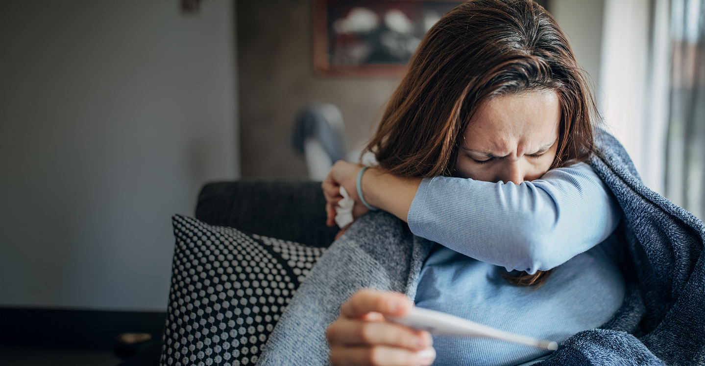 Sick woman coughing into arm while taking temperature.